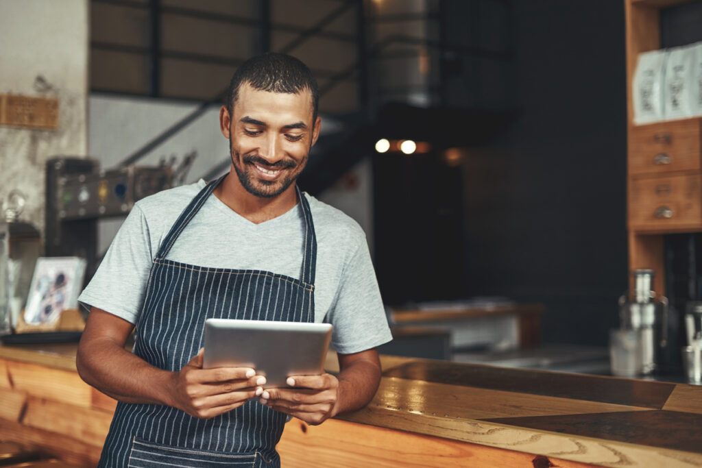 Portrait of a happy male owner looking at digital tablet while standing near the counter in his cafe